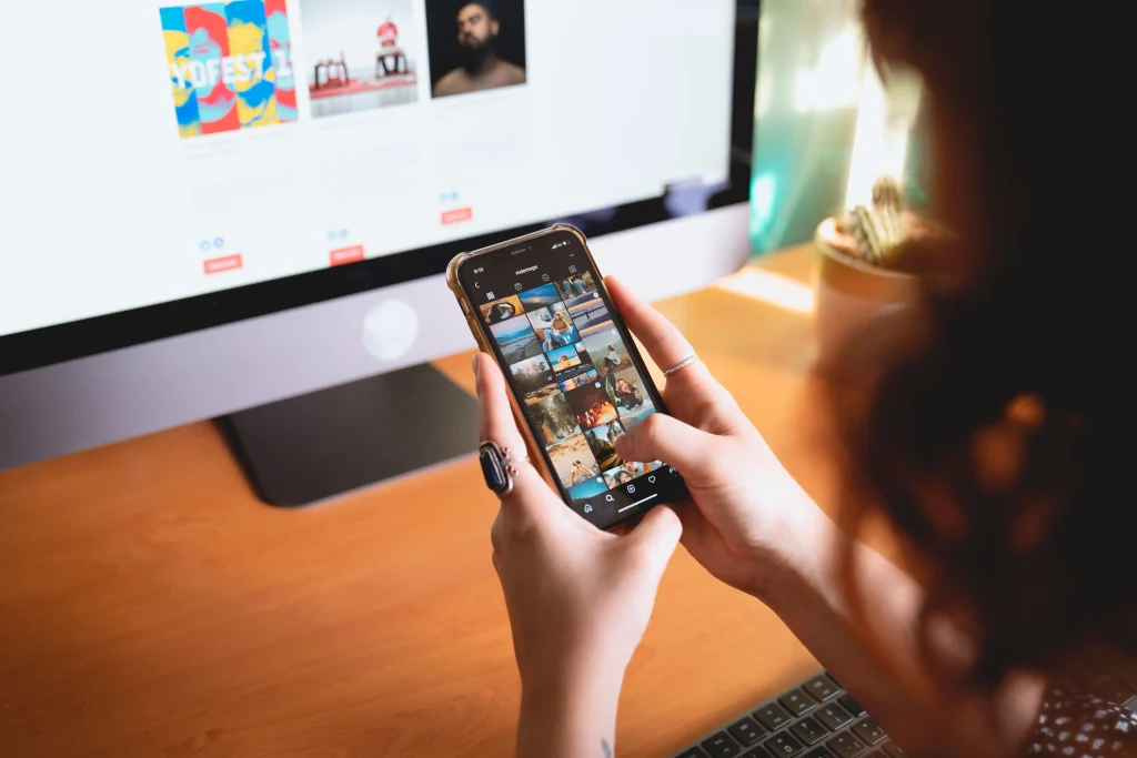 Social media manager parcourant des images sur un smartphone tout en étant assise à un bureau avec un écran d'ordinateur